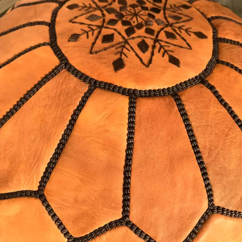 A tan leather pouf with dark embroidered stitching and a central geometric pattern.